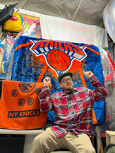 Justin Neely seated on a chair with a new york knicks fleece blanket hung over a clothing rack behind him